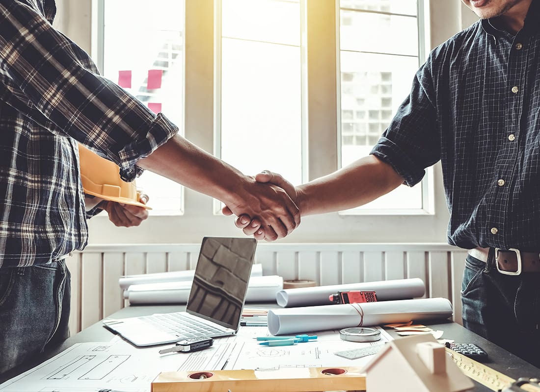 Insurance by Industry - Portrait of Two Contractors Shaking Hands Over a Table with Blueprints and a Laptop Inside a Bright Office